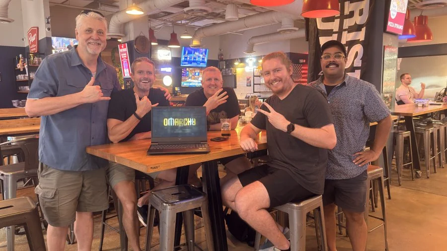 Five OATLUG founding members around a high-top table at Pour Taproom, a laptop in the middle showing the Omarchy logo, everyone throwing a shaka.